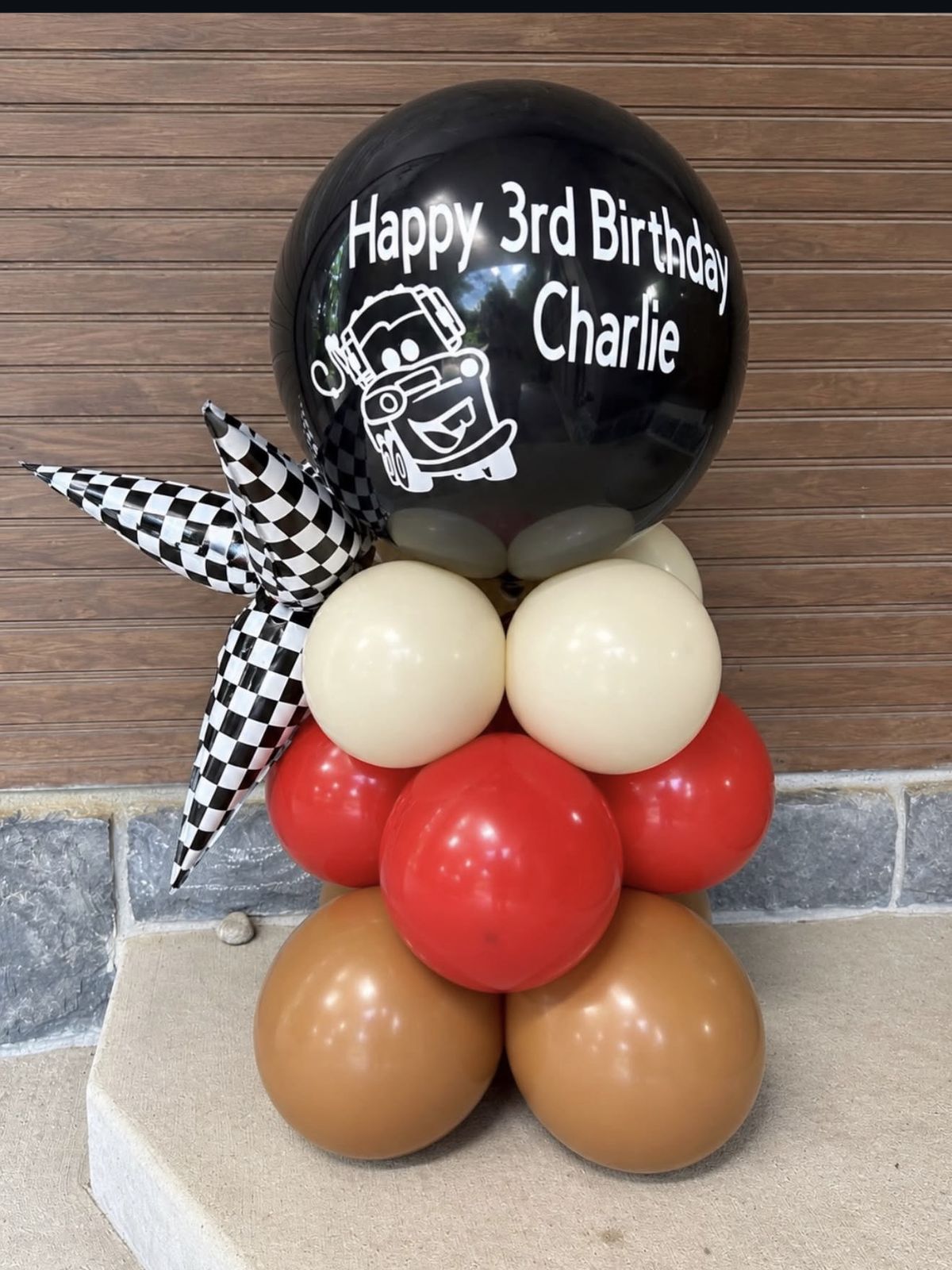 A grab-and-go balloon stack with a black personalized balloon reading Happy 3rd Birthday Charlie, a checkered flag balloon and red and tan balloons