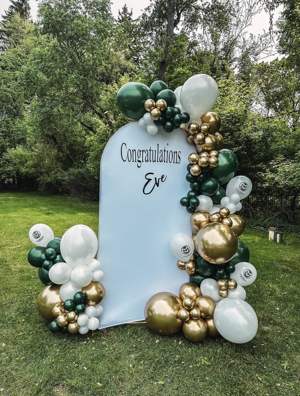 Forest green, chrome gold and white balloon garland on a light blue arch reading Congratulations Eve, outdoors on a lawn