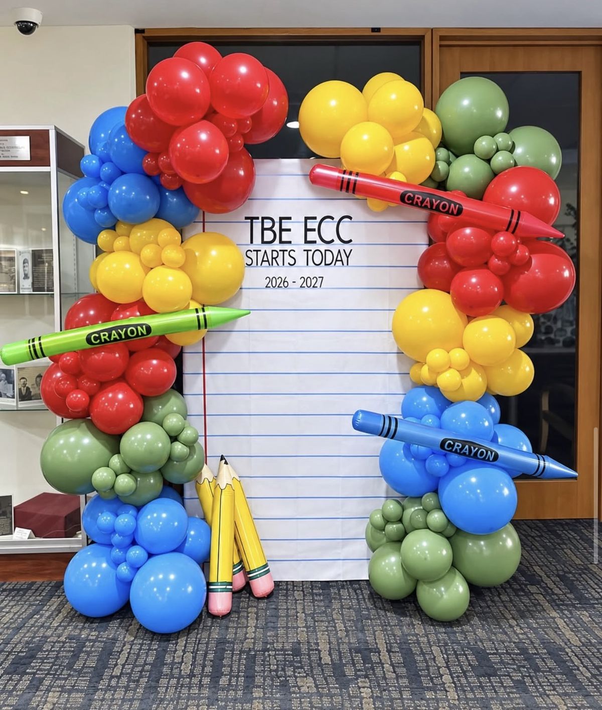Red, yellow, blue and green balloon garlands with giant crayon balloons around a notebook-paper backdrop for the first day of school