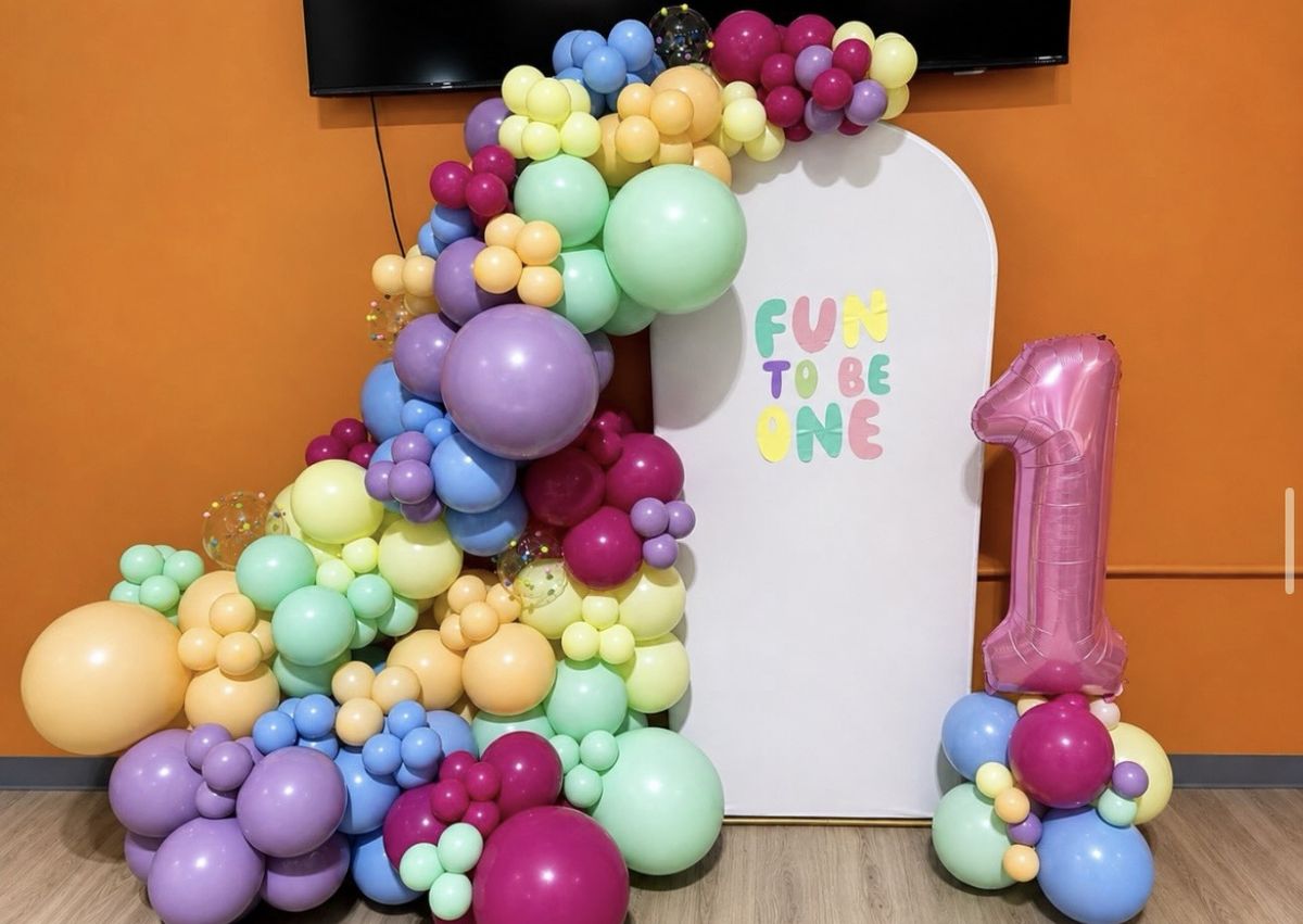 Pastel rainbow balloon garland in lavender, mint, yellow, peach, blue and magenta around a white arch reading Fun to be One with a pink number one balloon