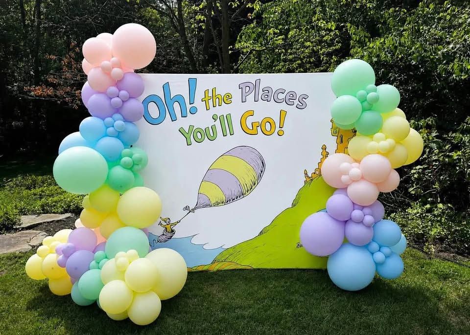 Pastel balloon garlands on both sides of an Oh the Places You'll Go backdrop in a garden