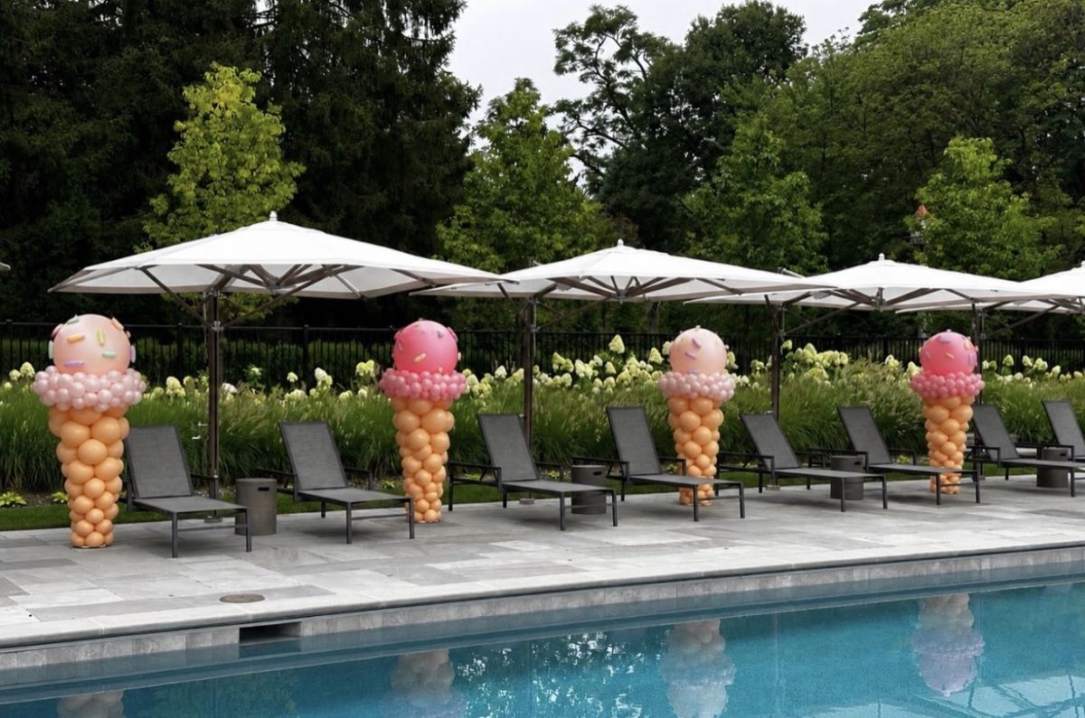 Four balloon towers shaped like ice cream cones with pink sprinkled scoops lined up beside a pool