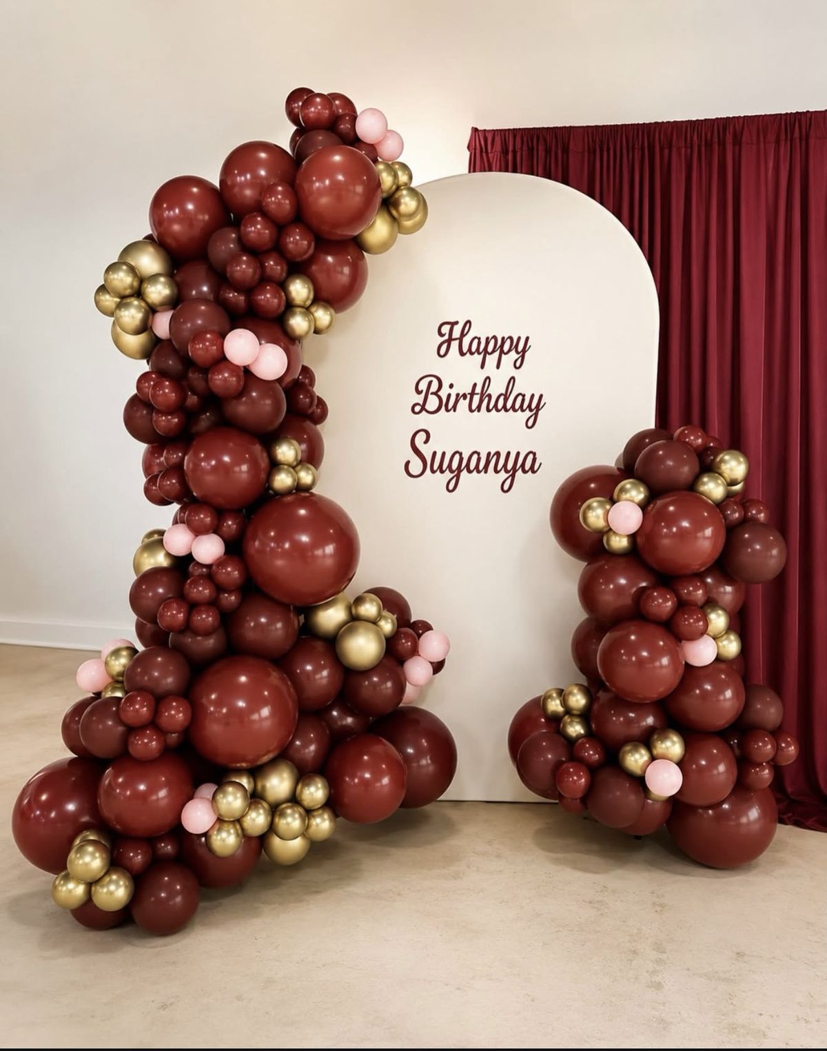 Burgundy, chrome gold and blush balloon garlands beside a cream arch that reads Happy Birthday Suganya
